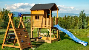 Une structure de jeux en bois, dotée d’un toboggan et d’un filet d’escalade, installée dans un espace verdoyant offrant une vue sur les montagnes lointaines.