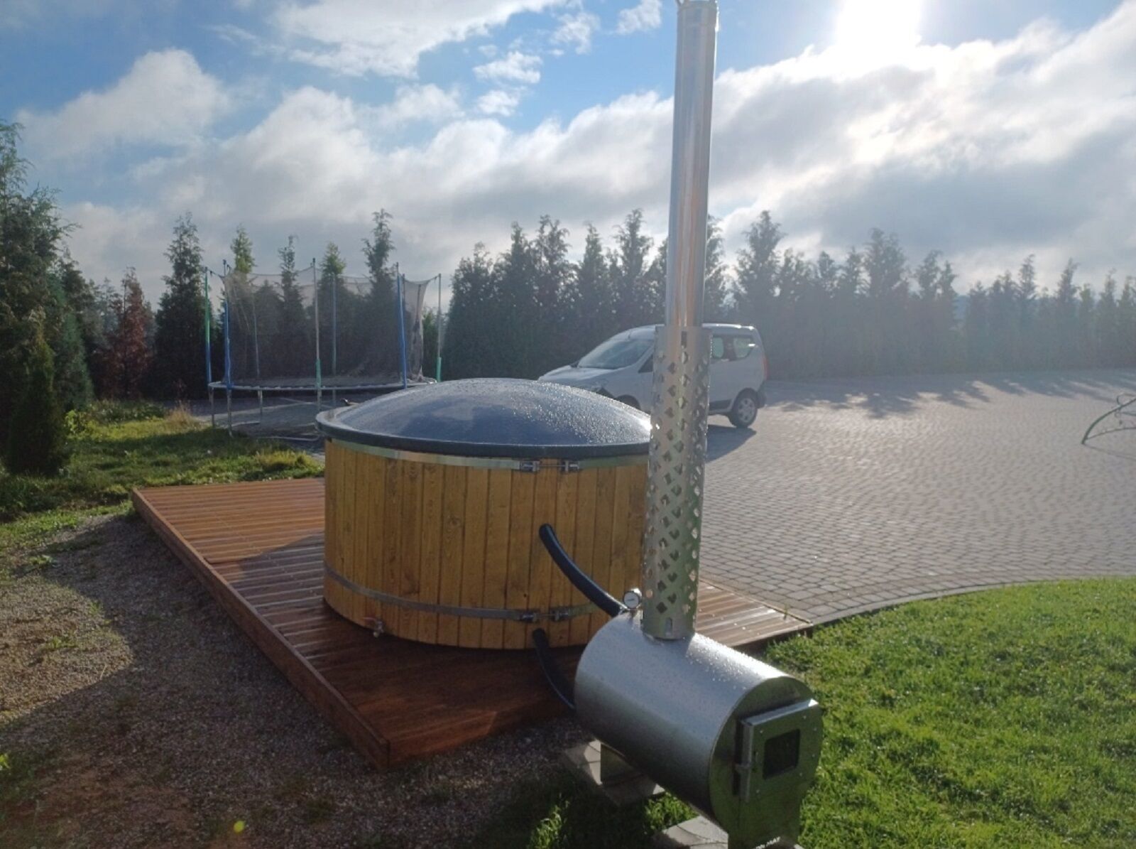 A sauna with a wooden deck and a metal chimney, surrounded by trees and a paved area with a car parked nearby.