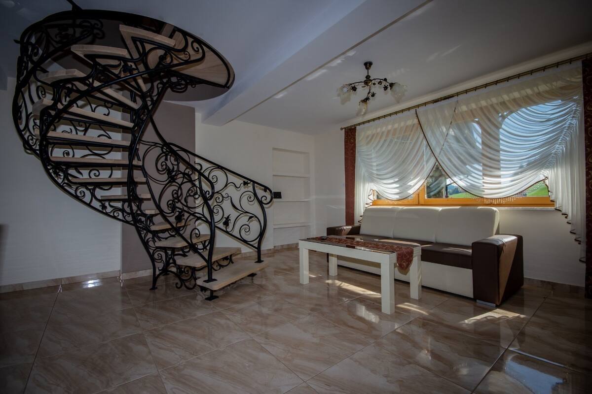 Un intérieur moderne avec un escalier en colimaçon, un canapé blanc et une grande fenêtre ornée de rideaux légers.