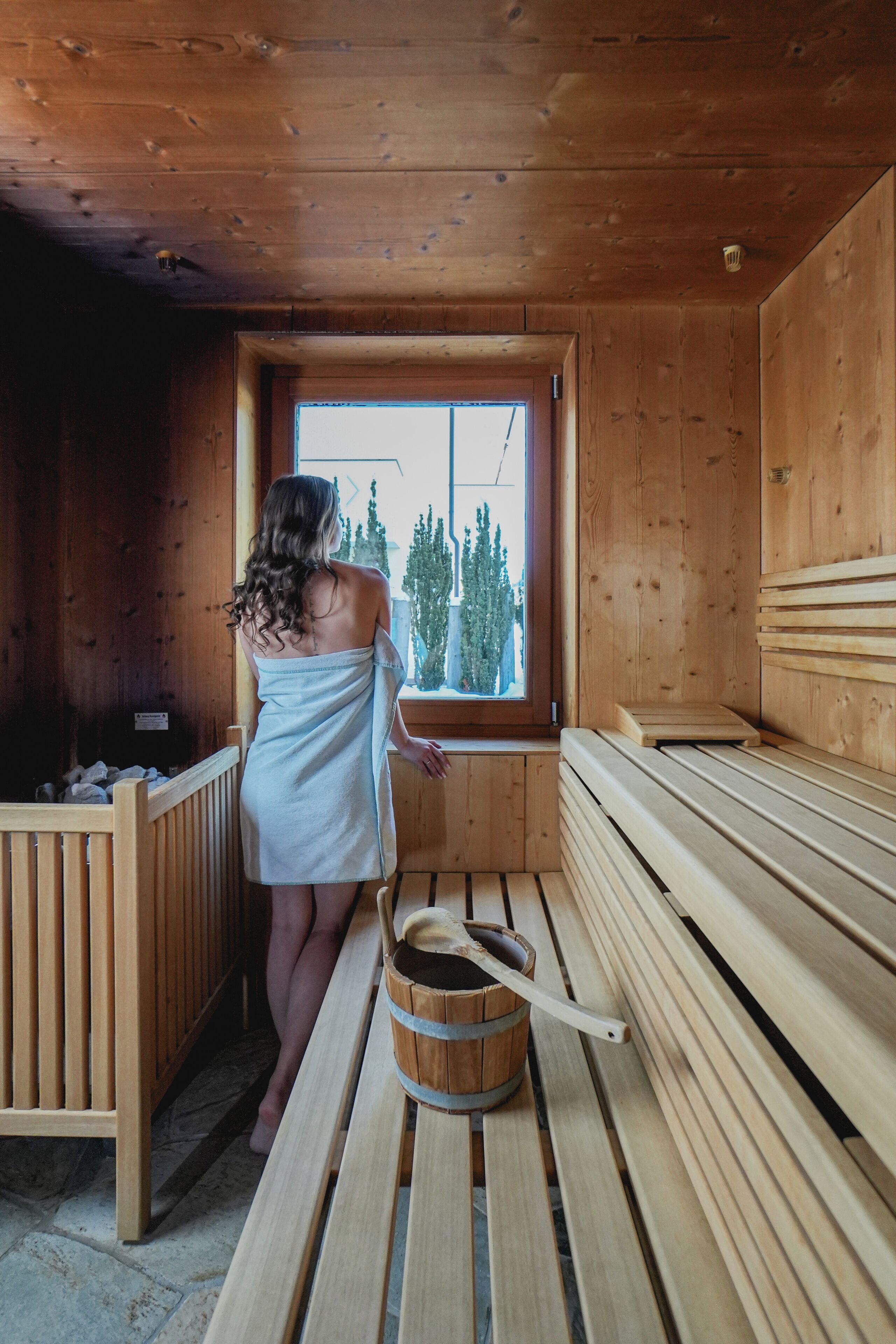 A sauna room with wooden benches, a wooden bucket, and a window with a view of trees.