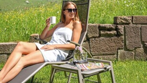 A person relaxing on a lounge chair with a magazine, holding a cup, in a garden setting with a mountain backdrop. - Hotel Bel Vert (Selva di Val Gardena)