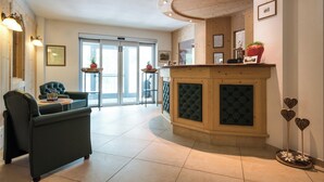 A reception area with a wooden counter, a green sofa, and a large window. - Hotel Bel Vert (Selva di Val Gardena)