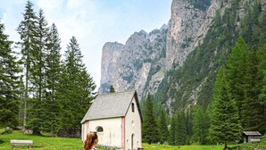 A person walking on a gravel path towards a small white building with a sloped roof, surrounded by green grass and trees, with mountains in the background. - Hotel Bel Vert (Selva di Val Gardena)