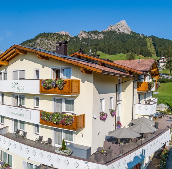 A hotel with a balcony with wooden railings and flower boxes, a terrace with outdoor seating, and a mountain view in the background. - Hotel Bel Vert (Selva di Val Gardena)
