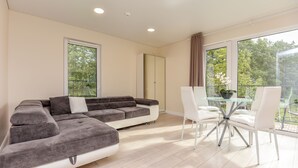 A modern living room with a sectional sofa, a glass-top dining table with white chairs, and large windows offering a view of trees. - Seaside apartments - Near Palanga Beach (Palanga)
