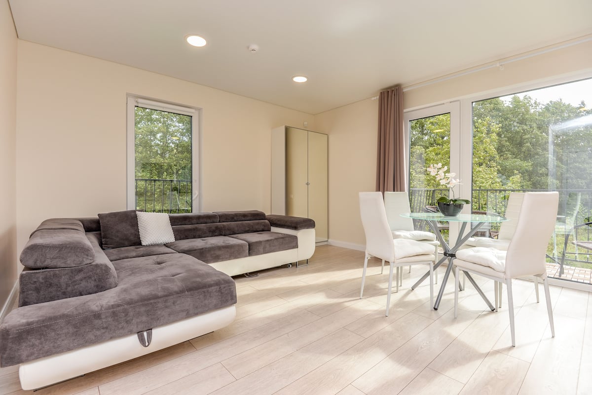 Een moderne woonkamer met een hoekbank, een eettafel met glazen blad en witte stoelen, en grote ramen met uitzicht op bomen.