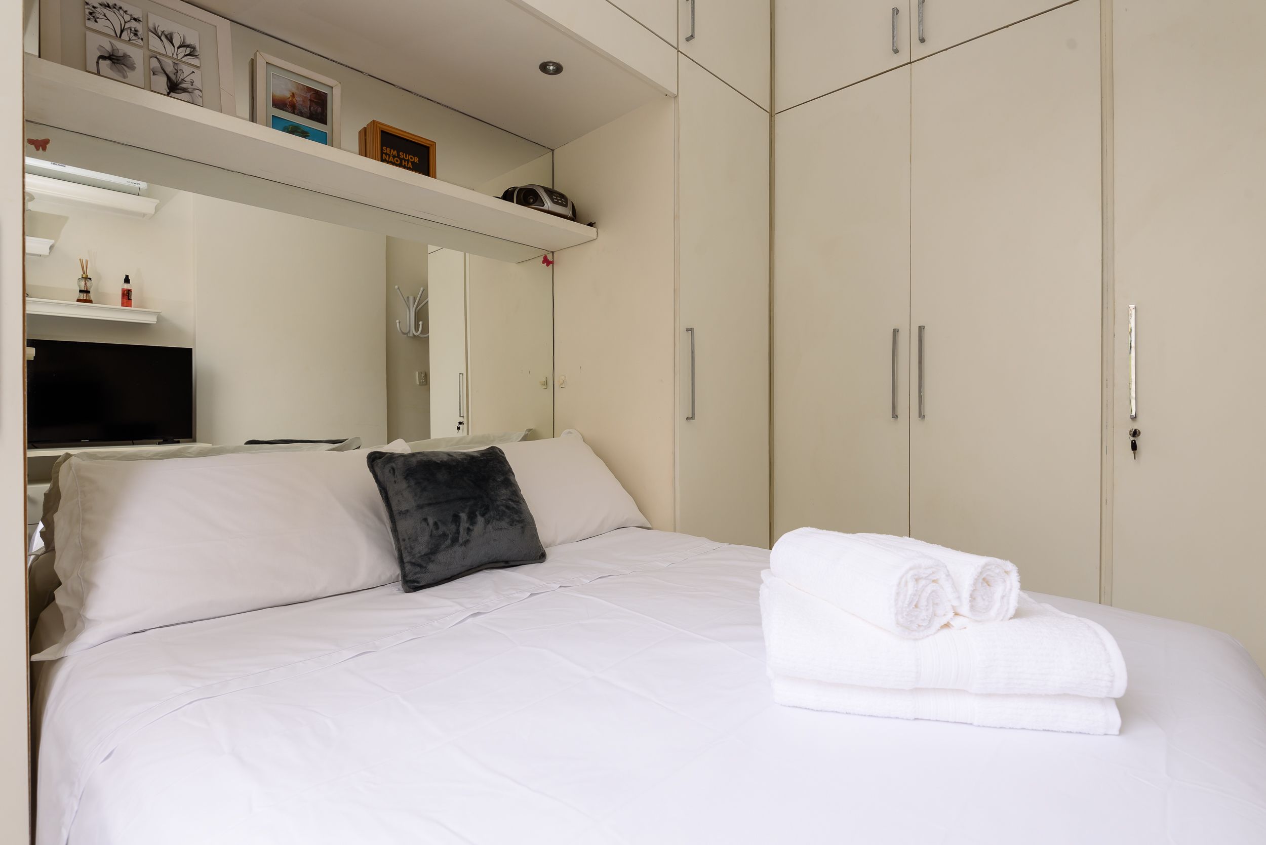 Een netjes opgemaakt bed met witte lakens en een zwart kussen, een opgevouwen handdoek aan de zijkant, en een televisie op een plank erboven.