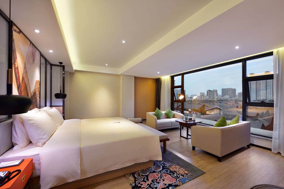 A modern hotel room with a large bed, a sofa, a small table, and a view of the cityscape through large windows.