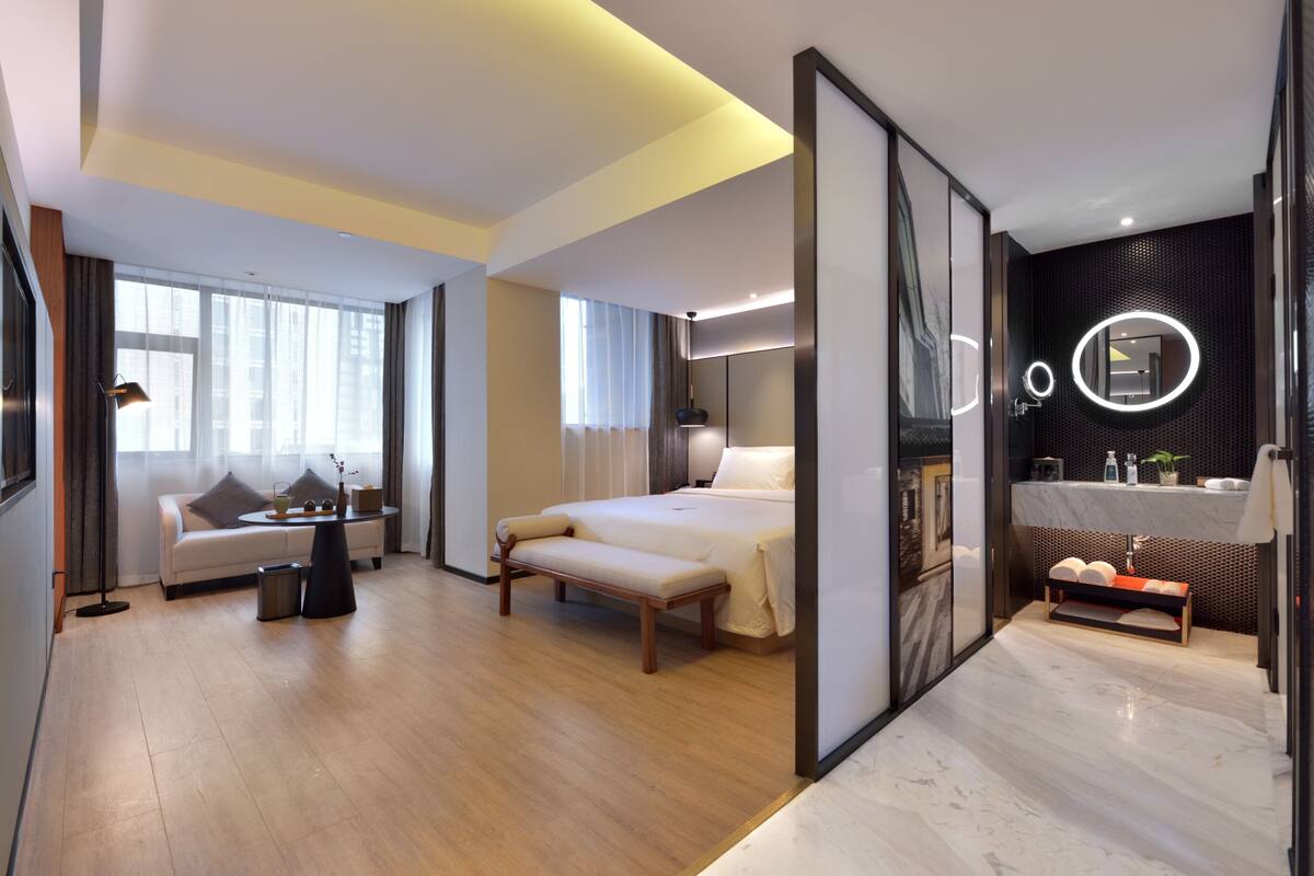 A modern hotel room with a large bed, a sofa, a bench, a desk, and a bathroom with a mirror and sink.