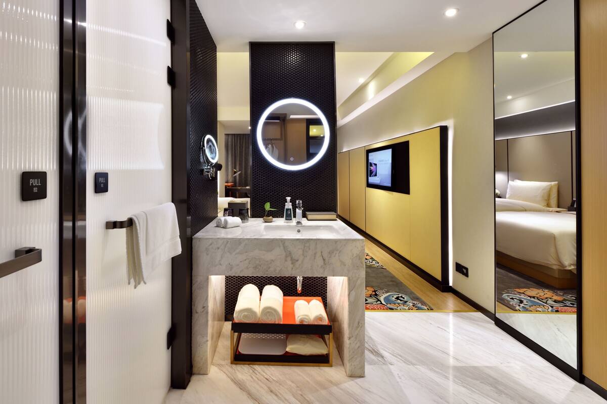 A modern hotel bathroom with a marble sink, a large mirror with a circular light, a toilet, and a shower area.