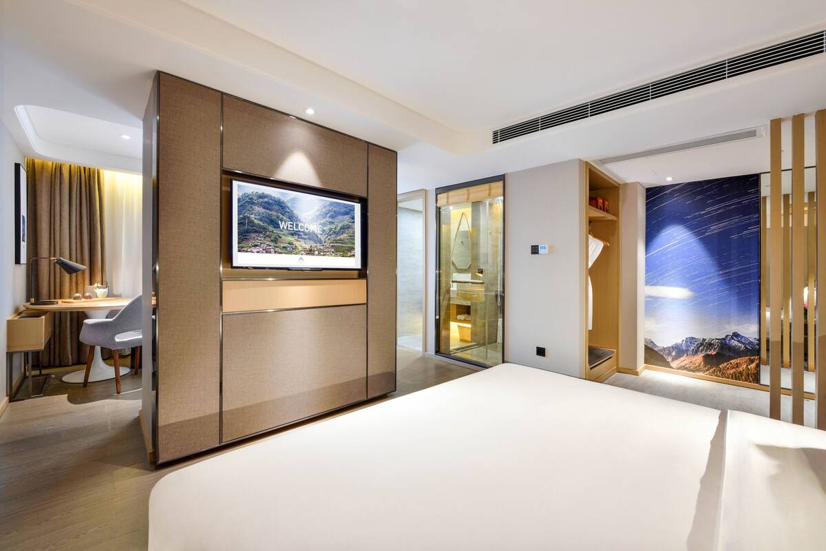 A modern hotel room with a large bed, a desk with a chair, a TV mounted on a wall, and a scenic view through a glass door.