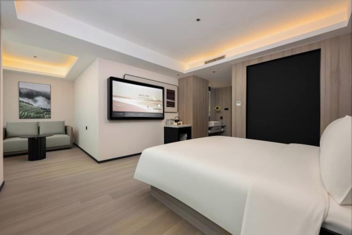 A modern hotel room with a large bed, a flat-screen TV mounted on the wall, a sofa, and a small table.