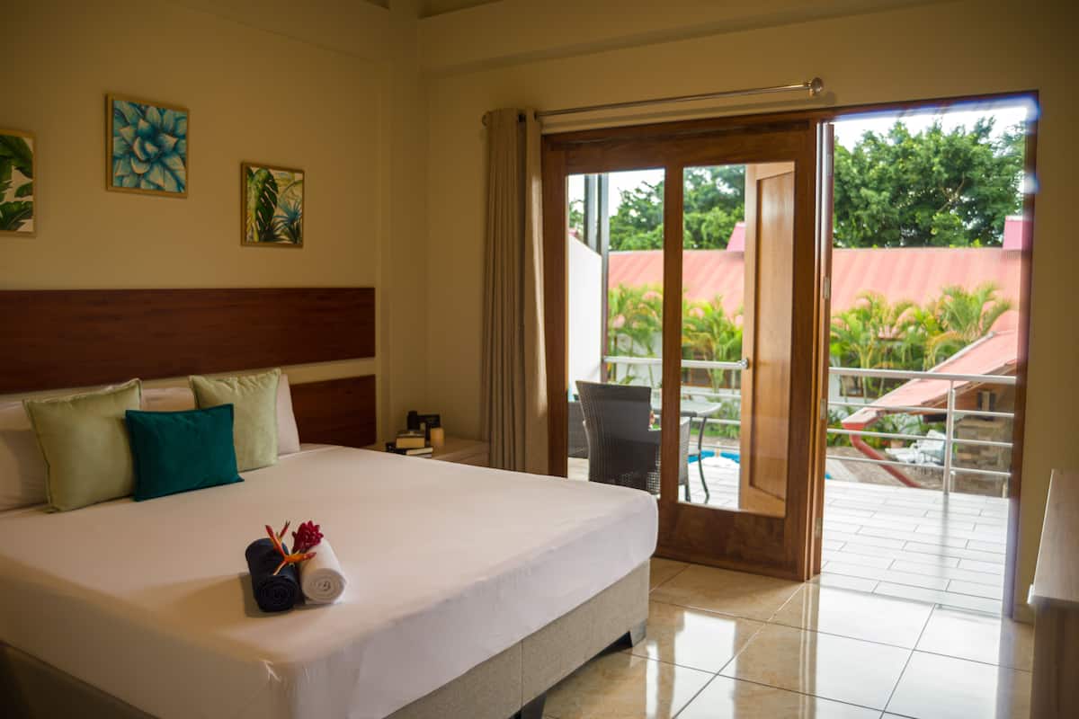 Une chambre d’hôtel avec un grand lit, une tête de lit en bois et une vue sur une piscine et la verdure environnante.