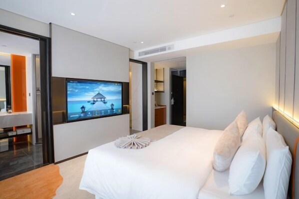 Quarto de hotel moderno com uma cama grande, televisão de tela plana na parede e vista para uma praia pela janela.