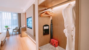 Ein modernes Hotelzimmer mit Holzboden, einer Sitzecke mit Tisch und Stühlen, einem Fernseher, einem Kleiderhaken mit Bademantel und einem kleinen roten Korb.