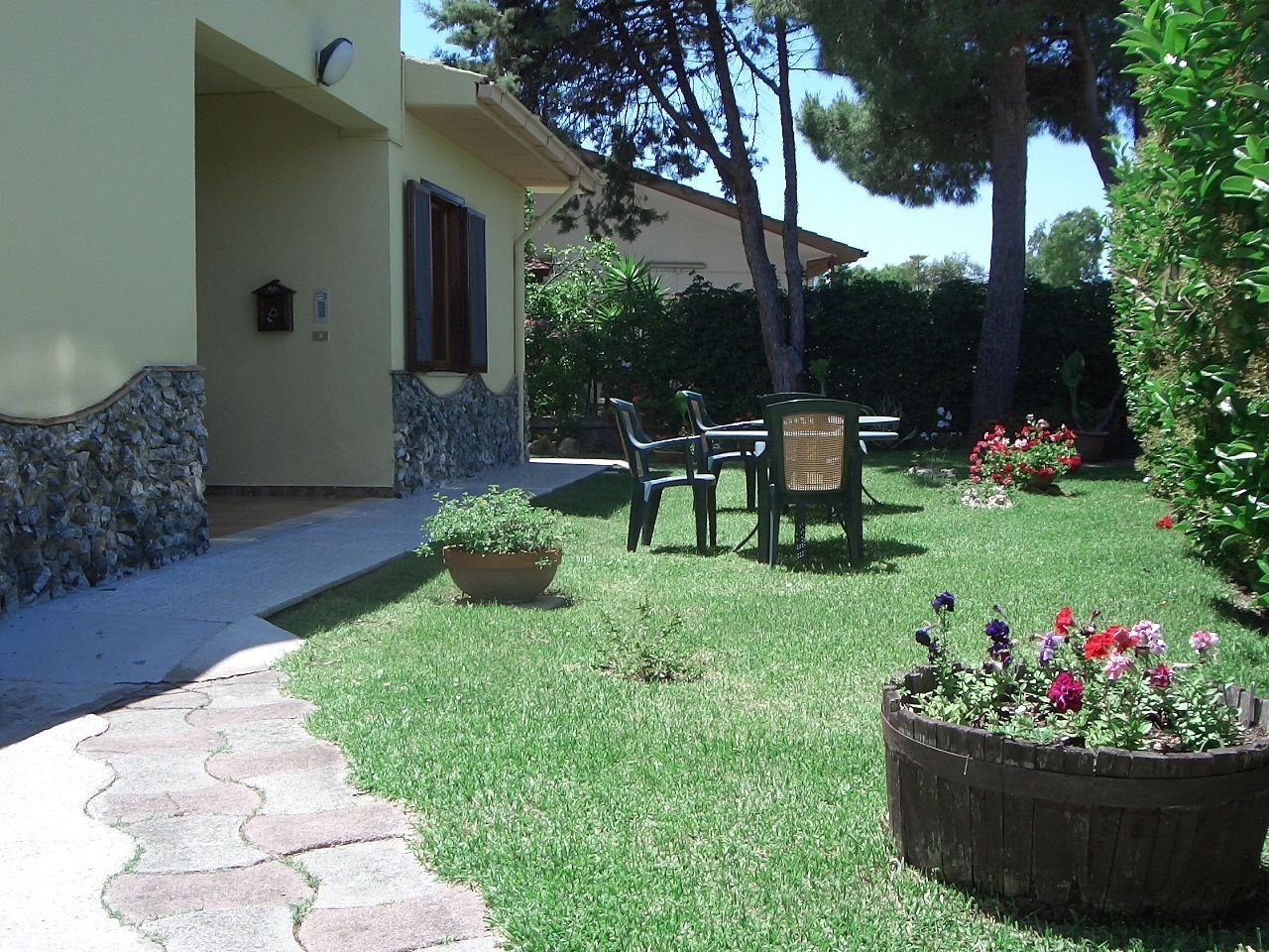 A house with a stone pathway leading to a lawn area with chairs and a table. There are potted plants and flowers in the garden.