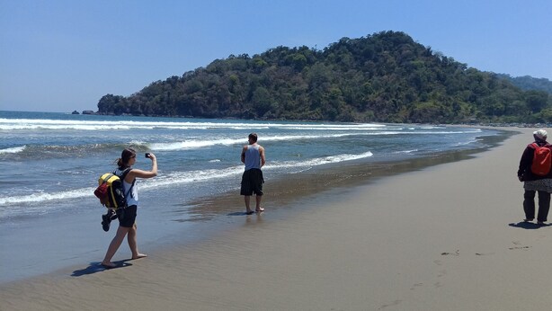 A beach with people walking along the shore, a forested hill in the background, and the ocean waves. - Gandrung City Hostel (Banyuwangi)