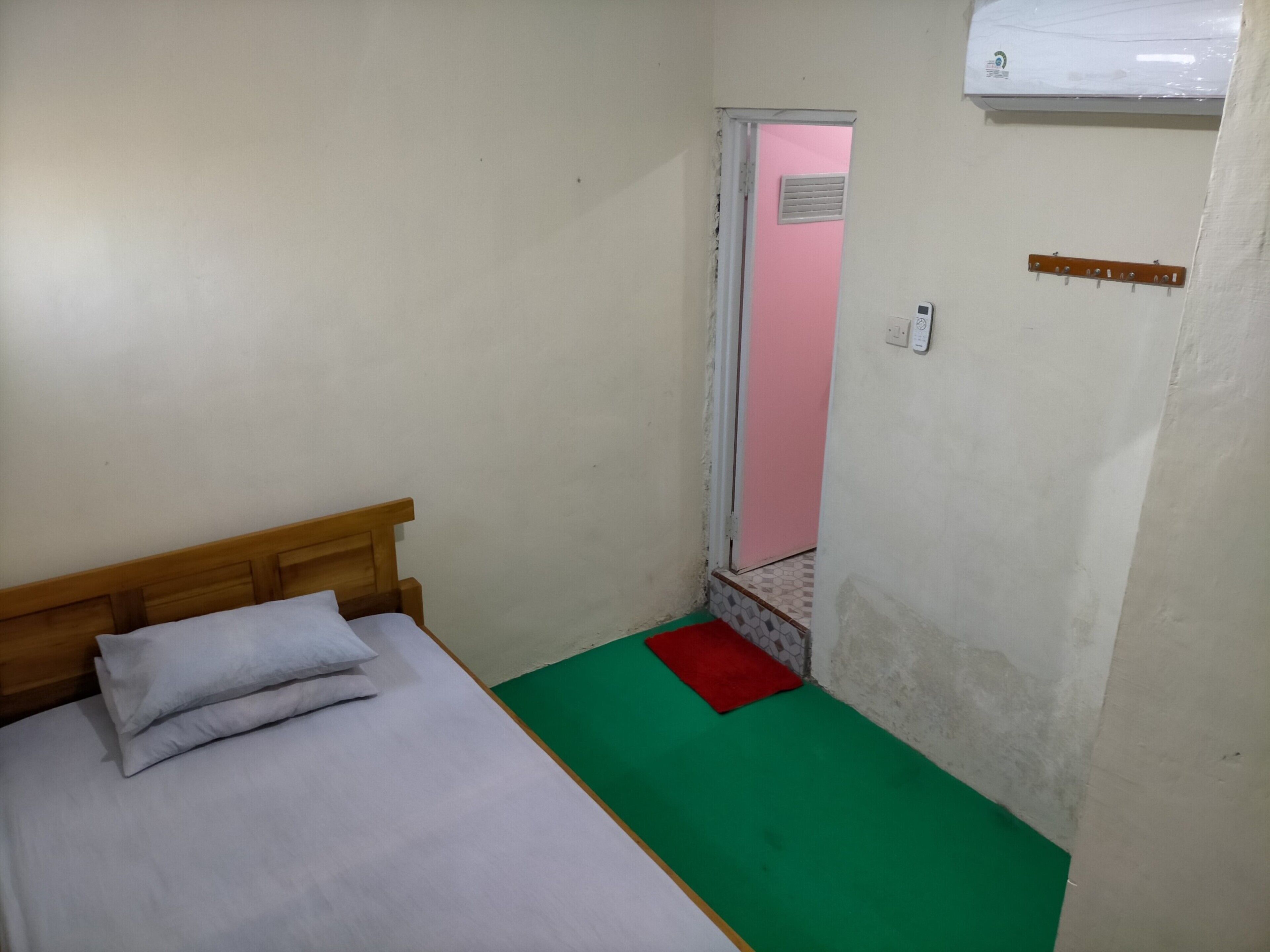 A small, single-bed room with a wooden headboard, a green and red rug, a pink door, and a white air conditioner on the wall.