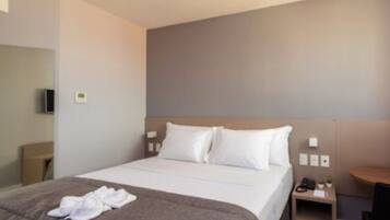 A hotel room with a bed, a bedside table with a lamp, a chair, and a small table with a phone and a cup.