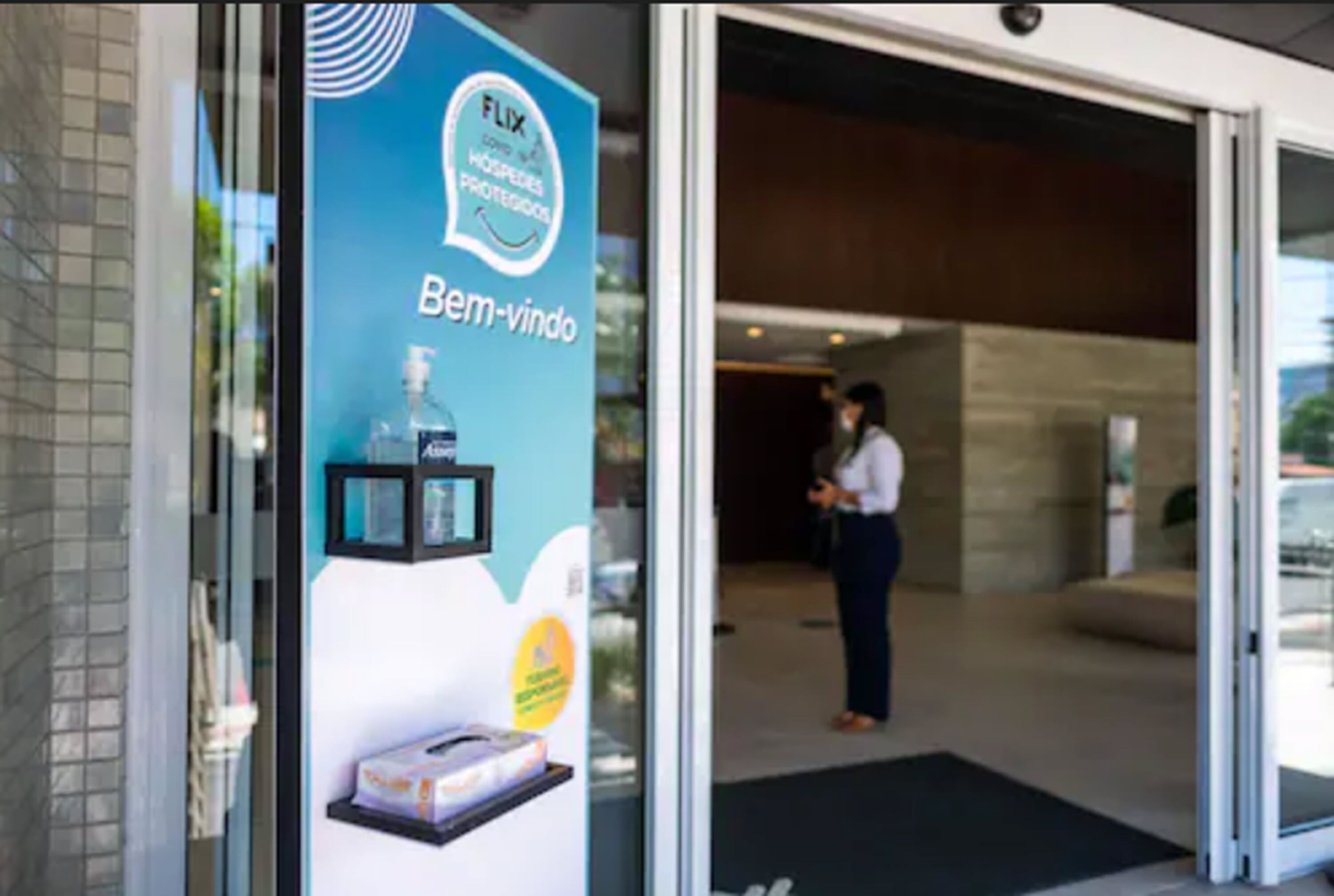 Entrada de un hotel con puerta de vidrio, un cartel que anuncia 'Bem-vindo' y 'FLIX', y una persona de pie en el interior.