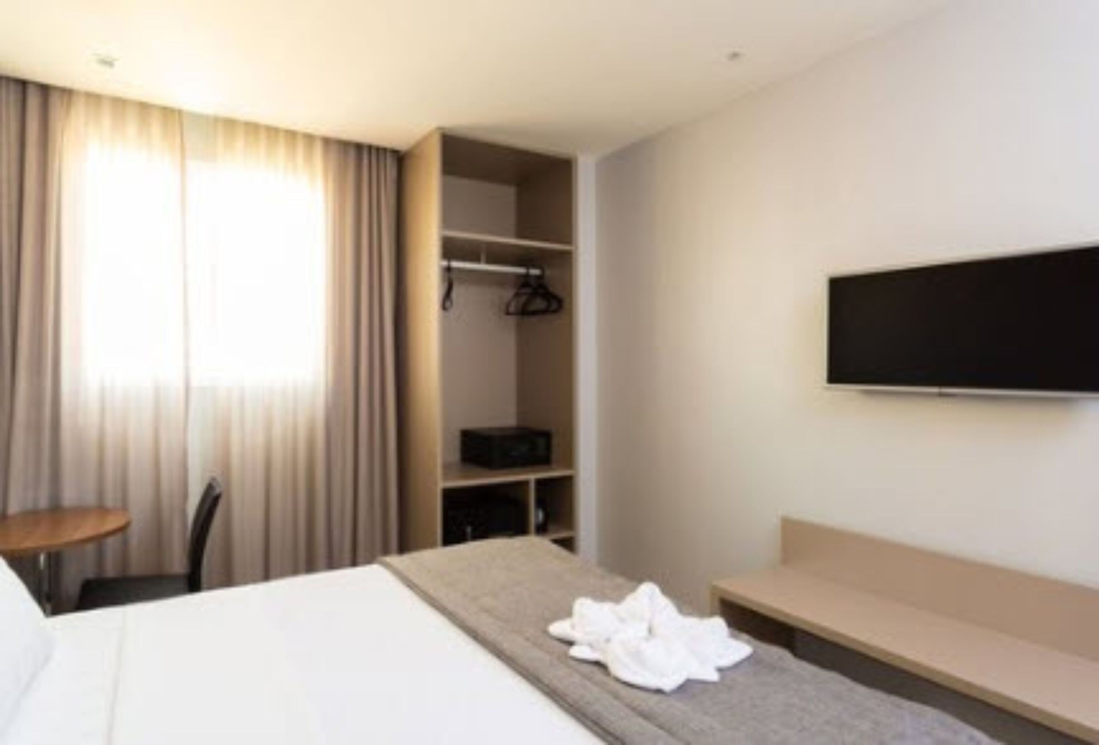 Una habitación de hotel con una cama, un televisor montado en la pared, una zona de estar y un armario con ropa colgada.