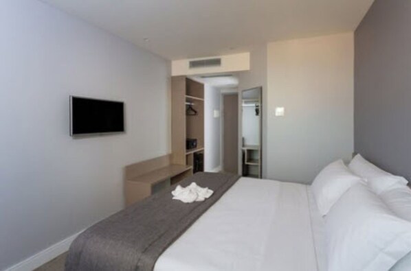 Quarto de hotel moderno com uma cama, televisão de tela plana na parede, closet espaçoso e banheiro visível através de uma porta aberta.