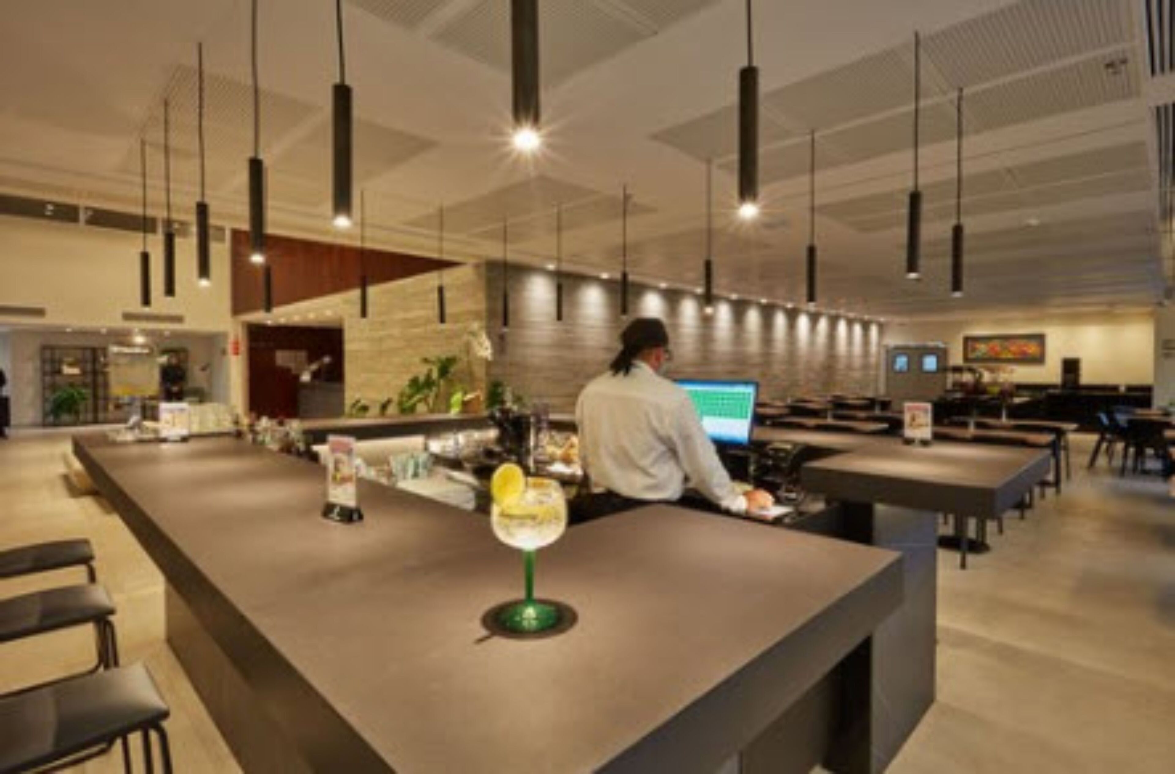 Zona de bar moderna con un bartender, una barra larga y lámparas colgantes.