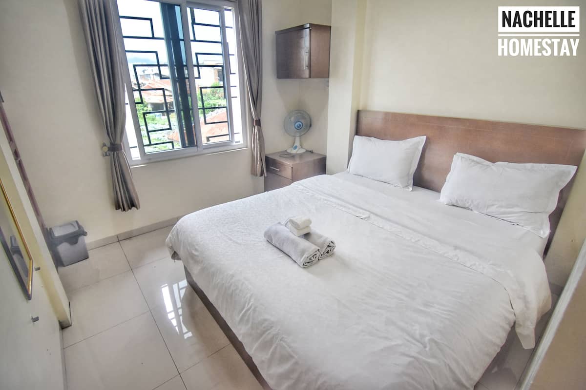 A neatly made bed with white linens and a rolled towel, a bedside table with a fan, and a window with curtains.