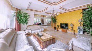 Una sala de estar amplia con un sofá grande, una mesa de centro y un televisor. Hay plantas en macetas y una pared de acento amarilla.