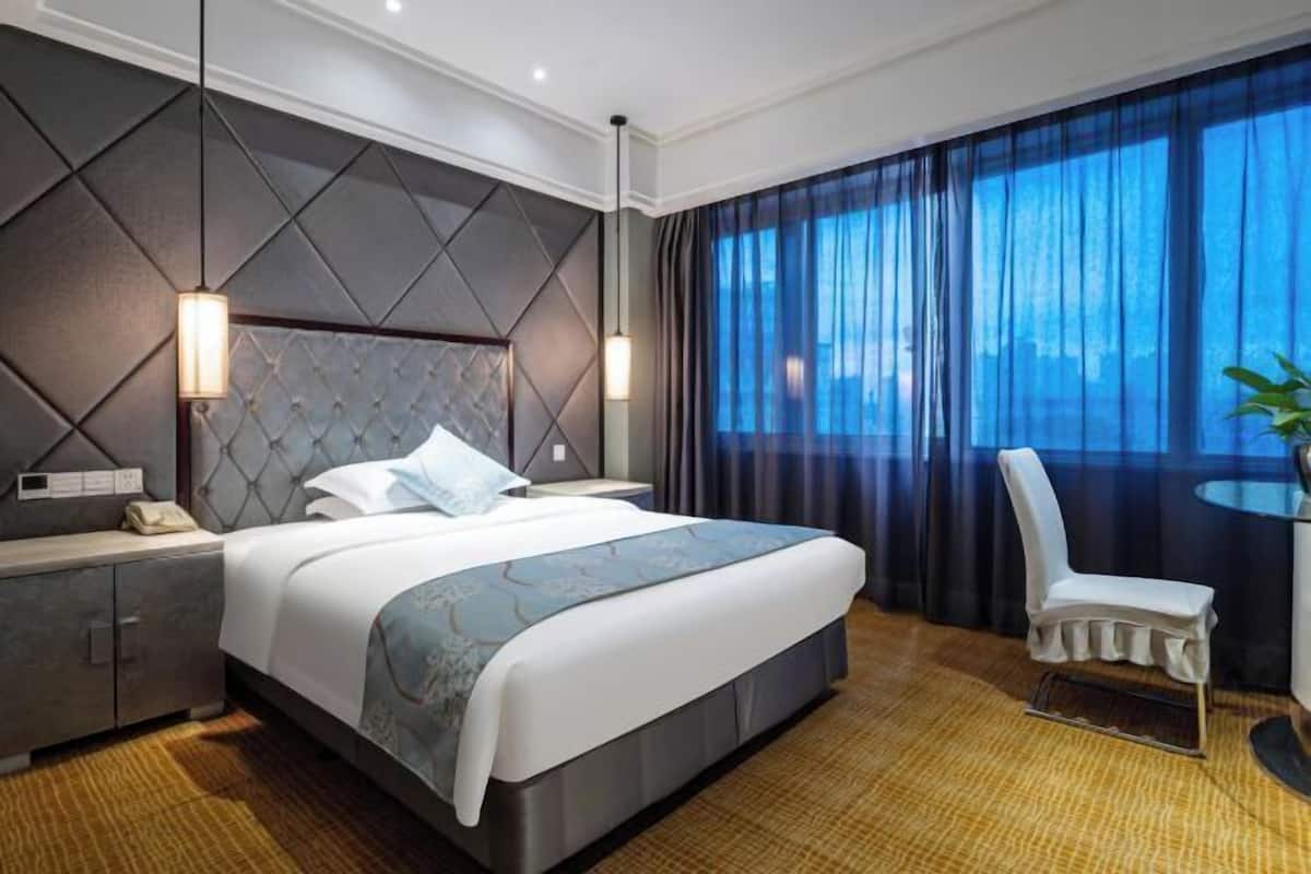 A modern hotel room with a large bed, a nightstand, a chair, and a view of the city through sheer curtains.