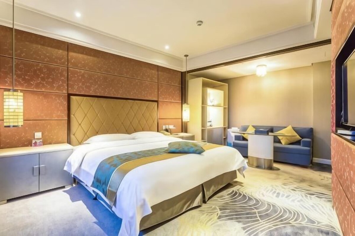 A hotel room with a large bed, a seating area with a sofa and armchairs, a TV, and a bathroom with a mirror and sink.