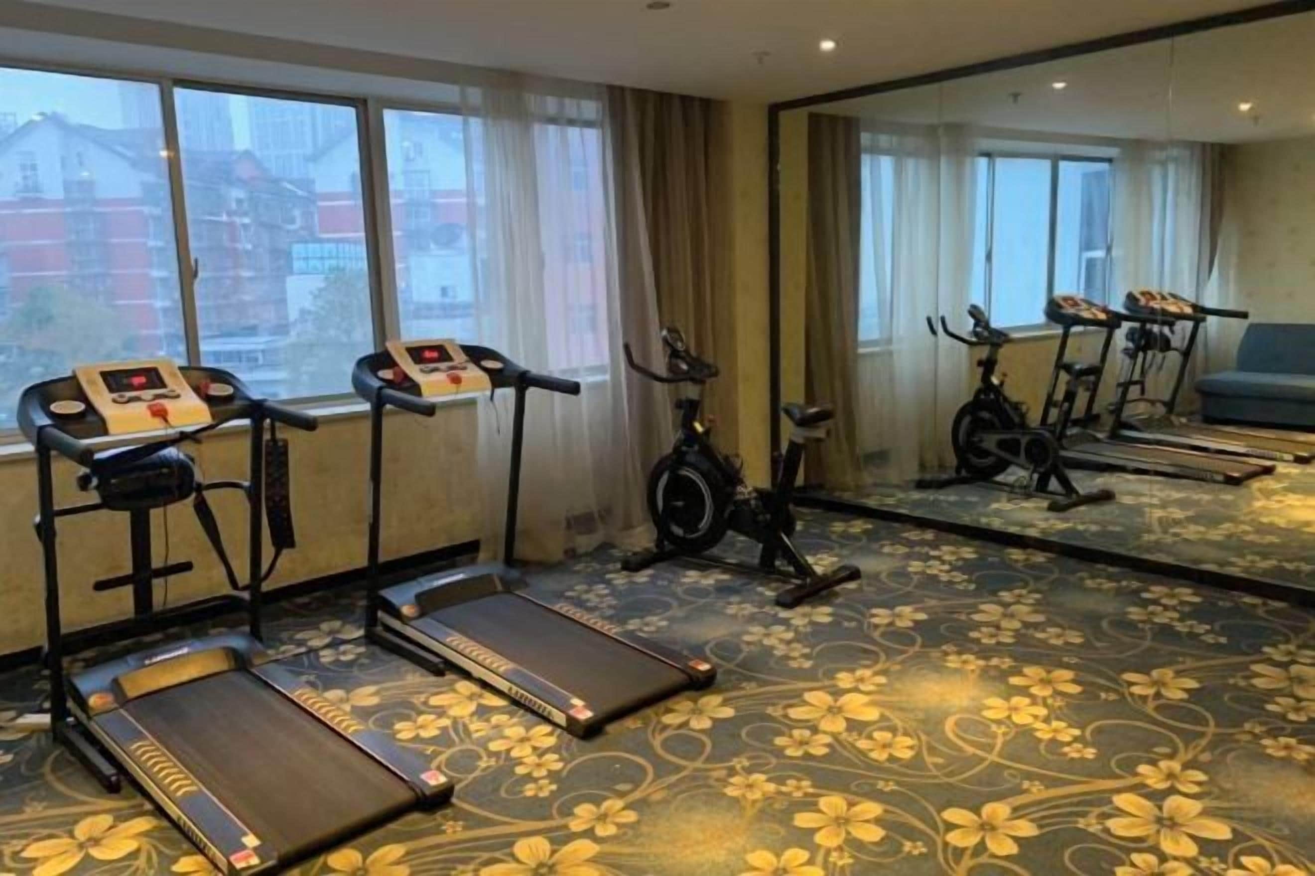 A hotel room with exercise equipment including treadmills and an elliptical, large windows, and a floral carpet.