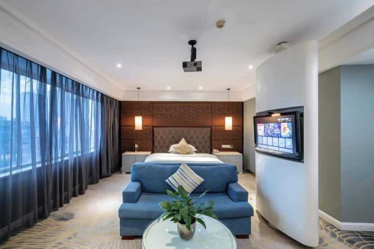 A modern hotel room with a large bed, a blue sofa, a glass coffee table with a plant, a TV mounted on the wall, and a view of the city through large windows.
