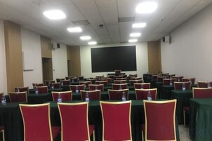 Un salón de conferencias con filas de mesas y sillas, una pantalla grande y un proyector.