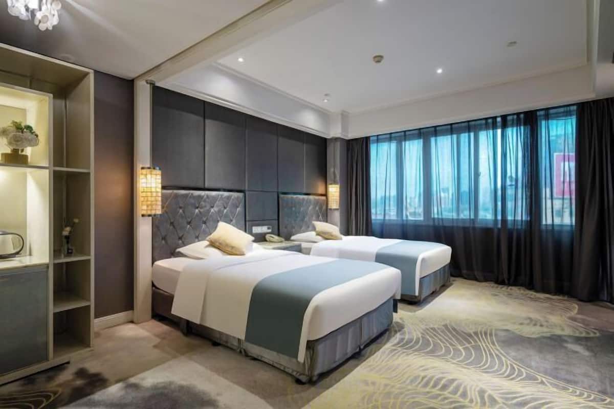 A hotel room with two beds, a large window with curtains, a built-in wardrobe, and a patterned carpet.
