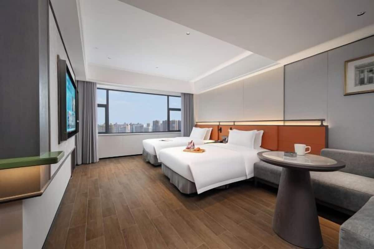 A modern hotel room with two beds, a sofa, a small table, and a large window offering a city view.
