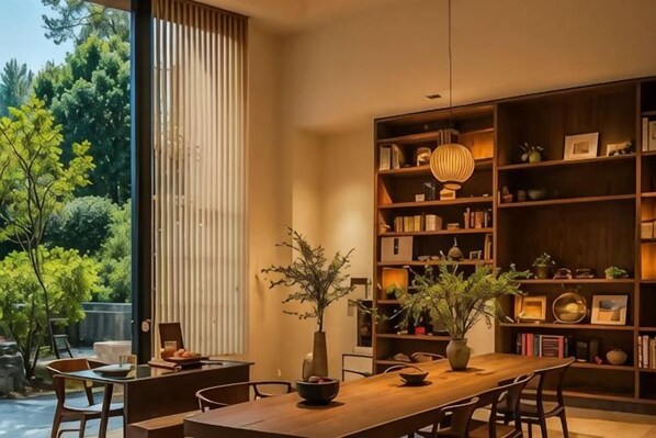 Un espace de repas moderne avec une table et des chaises en bois, une bibliothèque contenant divers objets et une vue sur la verdure à l’extérieur.