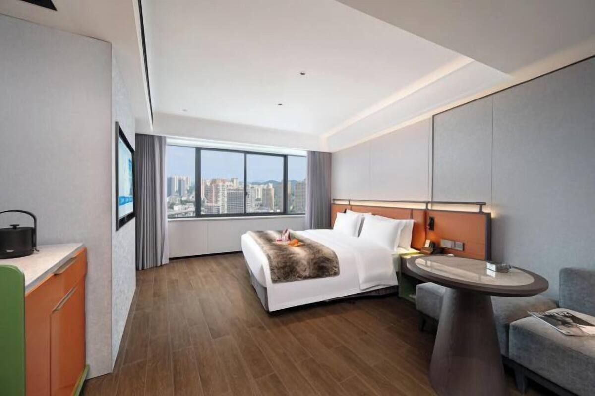 A modern hotel room with a large bed, a desk, a round table, and a view of the cityscape.