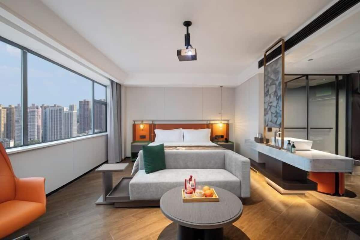 A modern hotel room with a large window, a bed with white linens, a gray sofa, a small round table with fruit, and a bathroom with a large mirror.
