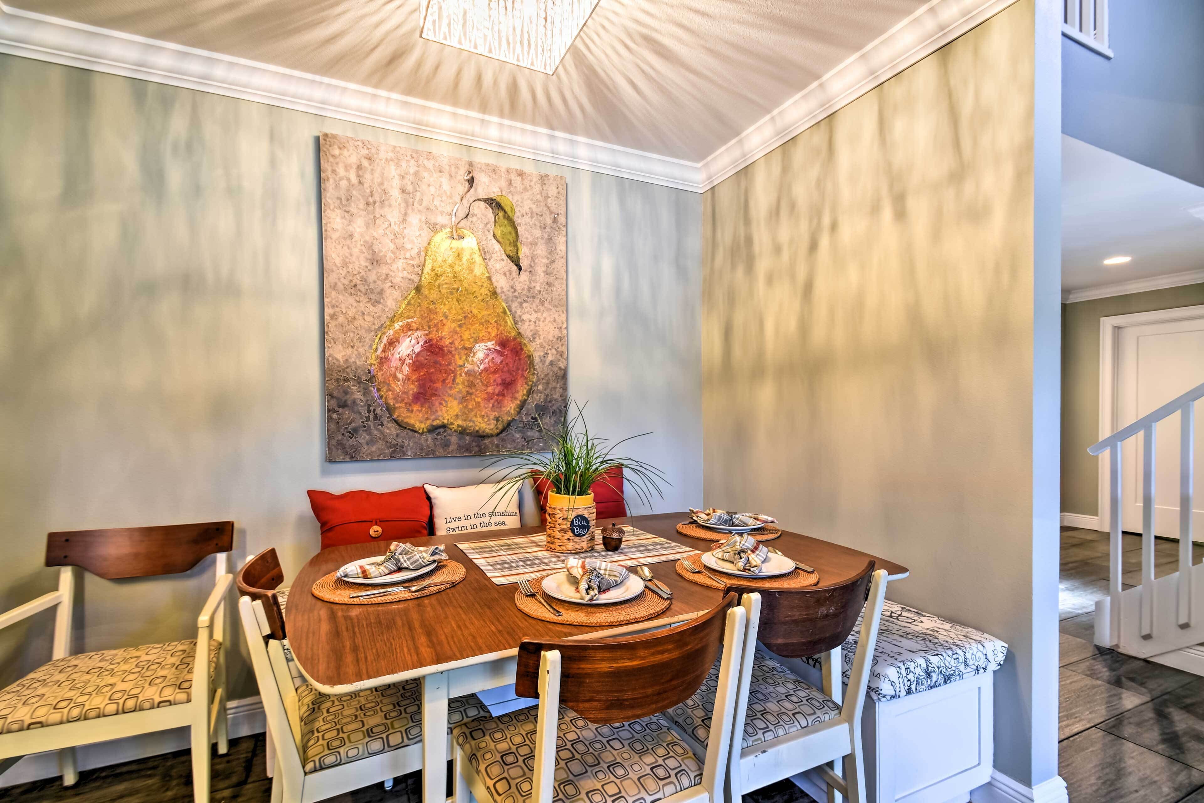 Een eetruimte met een houten tafel, gedekt voor vier personen, omringd door witte stoelen met gekleurde kussens. Daarboven hangt een groot schilderij van peren.