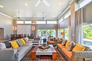 A spacious living area with a large sofa, a coffee table, and a dining table with chairs. There are two ceiling fans and large windows providing natural light. - Expansive St Pete Beach Home: Pool, Walk to Beach (St Pete Beach)
