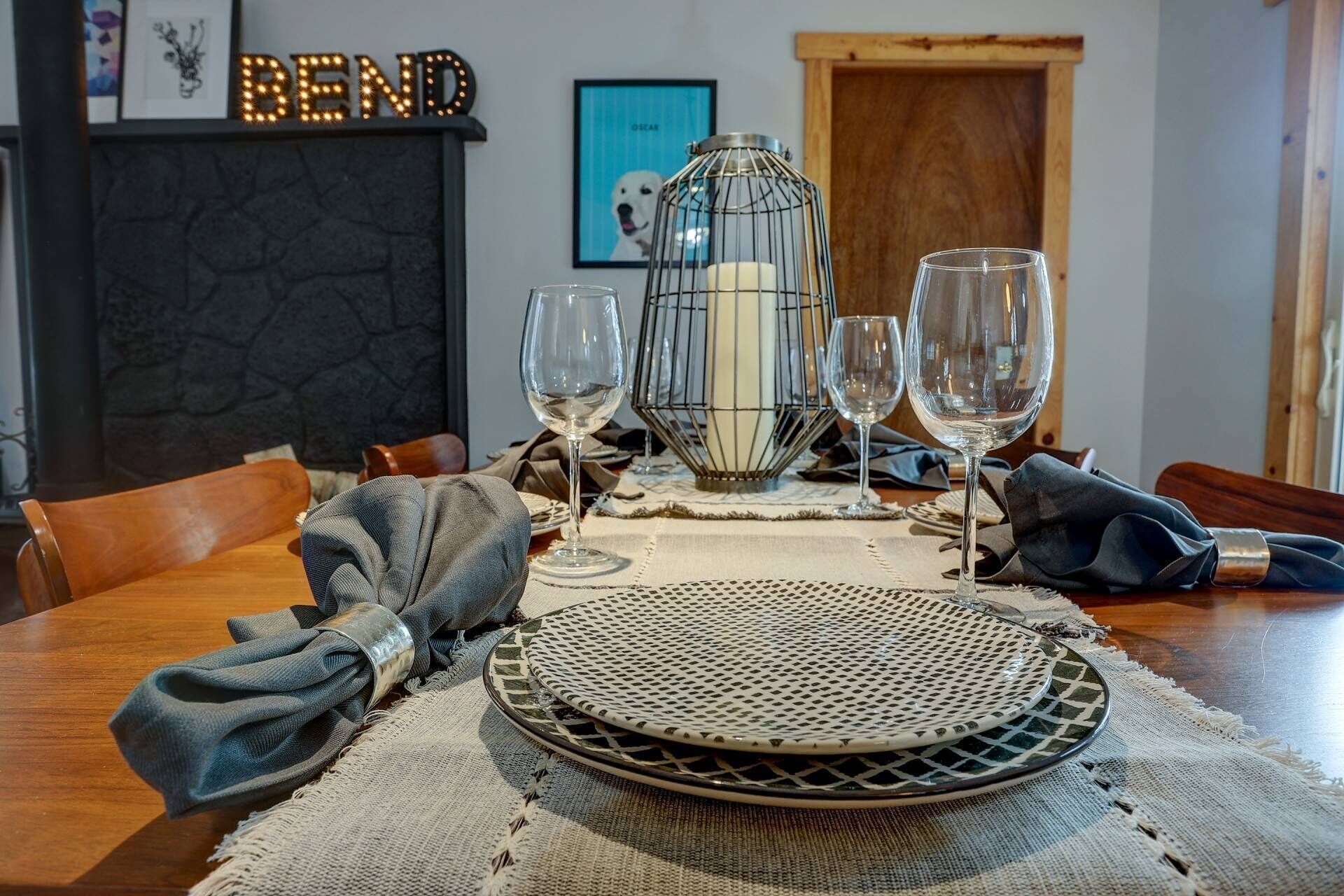 Een eettafel gedekt met borden, glazen en een kaars in een knus vertrek met een open haard en foto's in lijsten aan de muur.