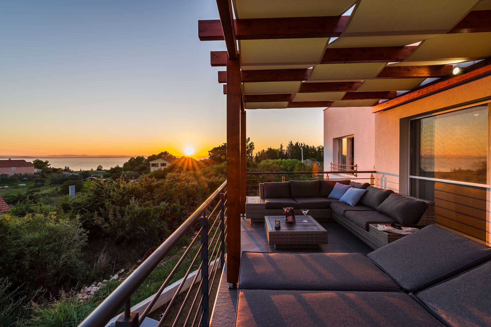 Un balcon avec un canapé et une table basse, d’où l’on peut admirer un coucher de soleil sur une étendue d’eau.