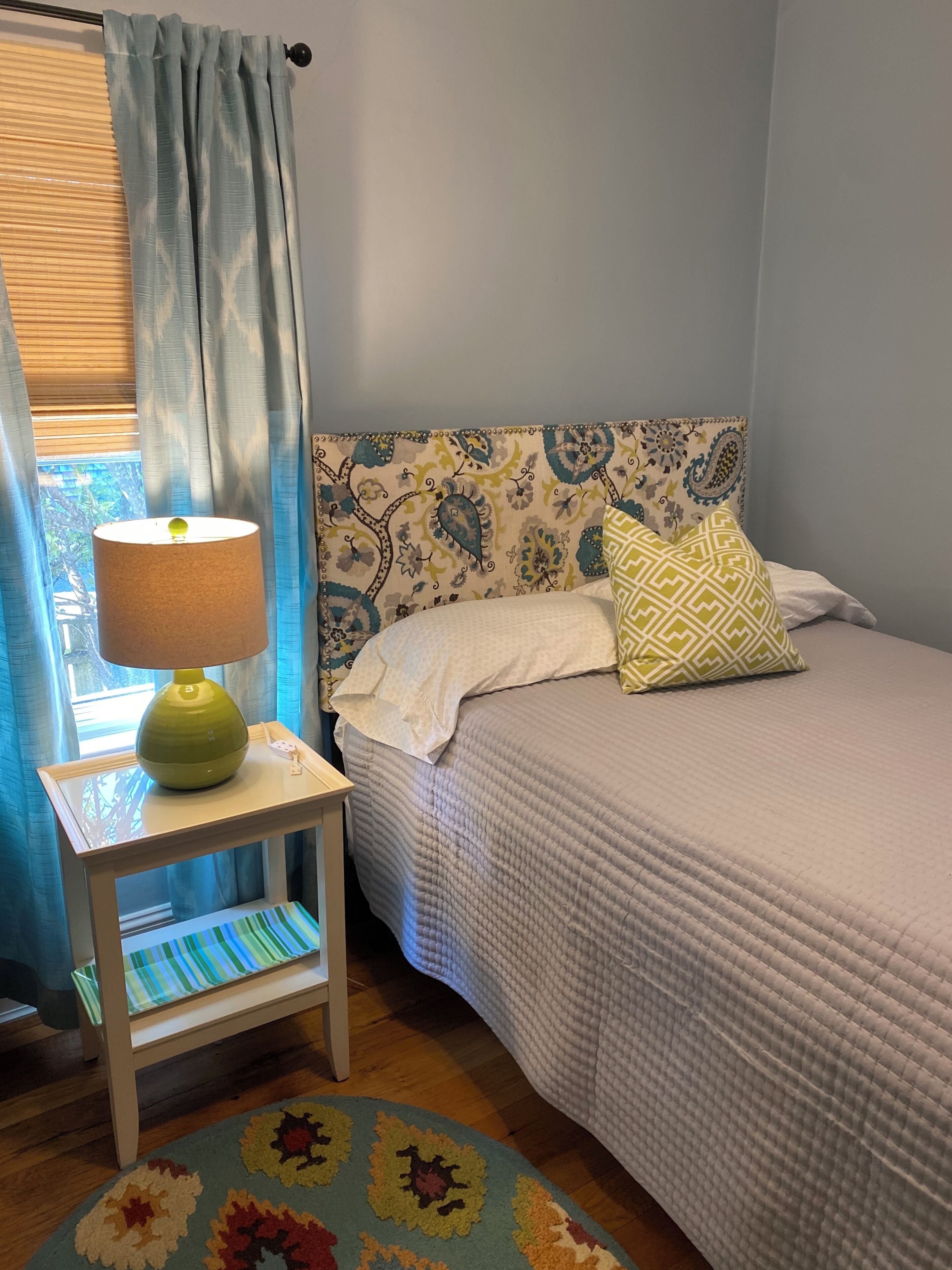 A bedroom with a bed, a bedside table with a lamp, a patterned headboard, and a window with curtains.