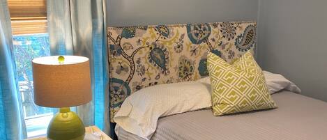 A bedroom with a bed, a bedside table with a lamp, a patterned headboard, and a window with curtains.