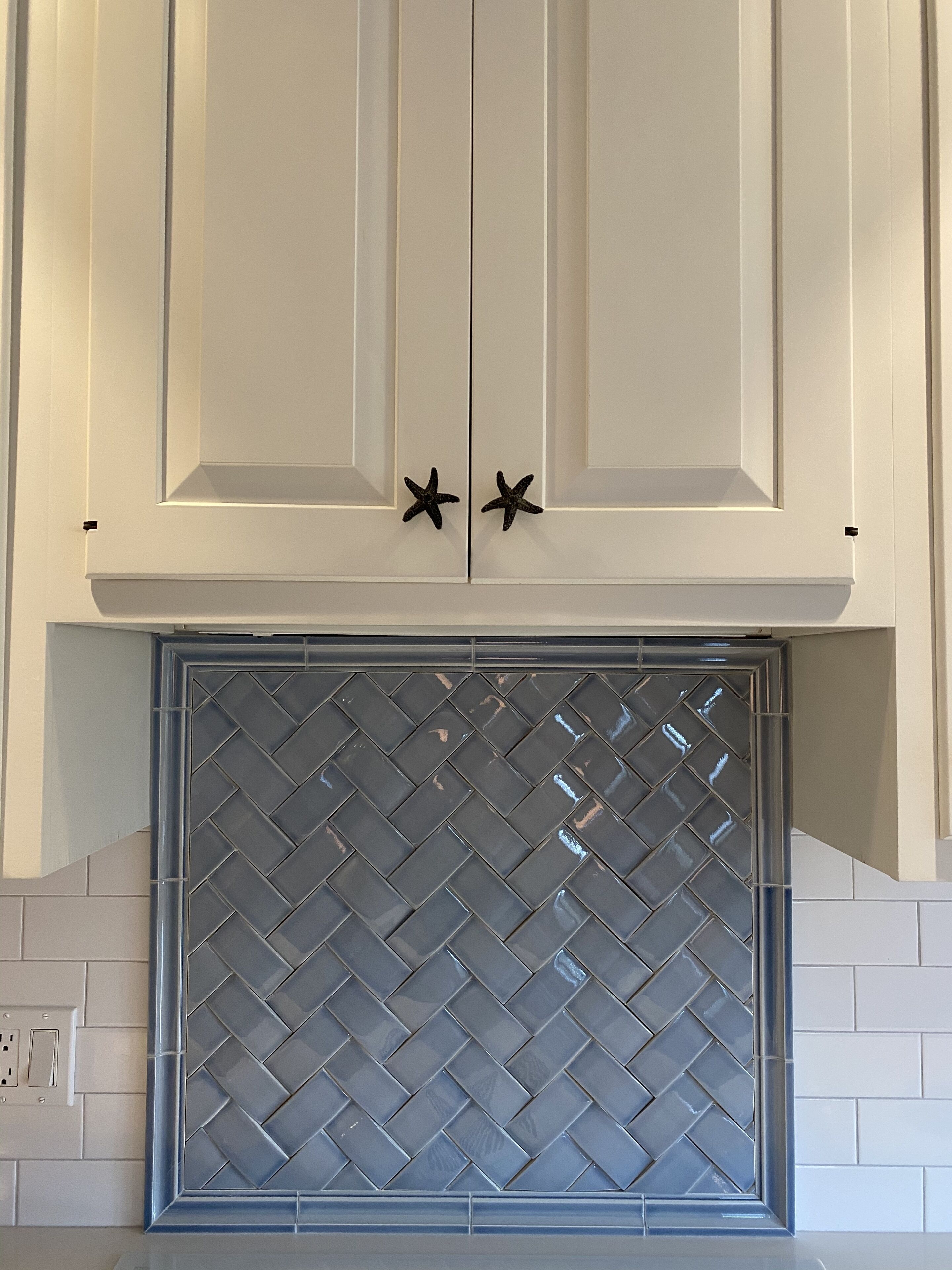 Cucina con mobili bianchi, zoccolo a motivi a spina di pesce e due pomelli a forma di stella marina.