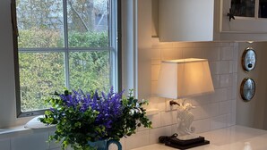 Una cocina con una ventana, un jarrón azul con flores moradas, una lámpara blanca y un caballito de mar decorativo.