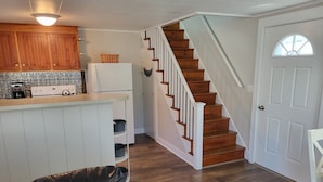 A modern kitchen with wooden cabinets, a white refrigerator, and a built-in oven. There is a staircase with wooden steps leading to a door with a window. - Cozy Cottage at Footbridge Beach - Great Fall and Spring Rates (Ogunquit)
