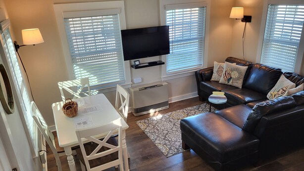 A modern living room with a flat-screen TV, a black leather sofa, a white dining table, and a small side table. - Cozy Cottage at Footbridge Beach - Great Fall and Spring Rates (Ogunquit)
