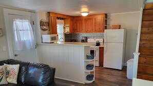 A compact kitchen with wooden cabinets, a white refrigerator, a microwave, and a stove. There is a small island with shelves and a white chair. - Cozy Cottage at Footbridge Beach - Great Fall and Spring Rates (Ogunquit)
